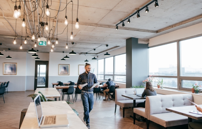 A man walking through an office with people sitting at tables.